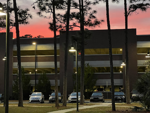 Streaks of pink, orange, and cotton candy blue take over the clouds as the sun sets behind cars parked in front of the Fine Arts Center Parking Garage (Building 44A)