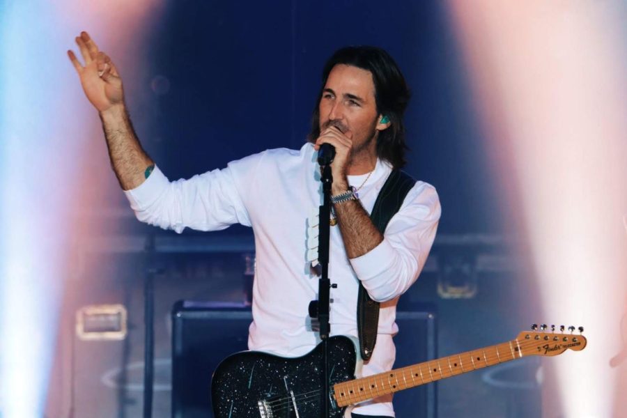 Country artist Jake Owen holds a guitar pick in the air with one hand while the other holds a microphone during a concert