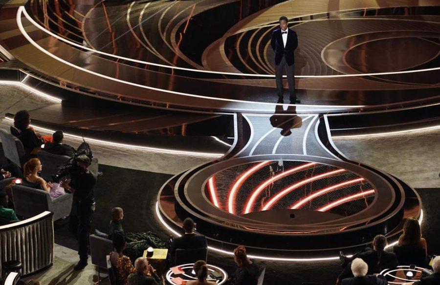 Presenter Chris Rock speaks onstage as Will Smith and Jada Pinkett, bottom left, listen from their seats at the Oscars, Sunday, March 27, 2022, at the Dolby Theatre in Los Angeles.