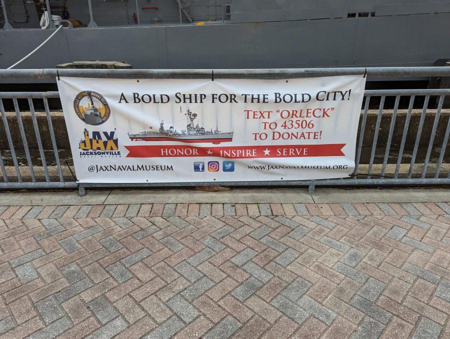 A sign from JHNSA placed along the riverwalk with the USS Orleck,
