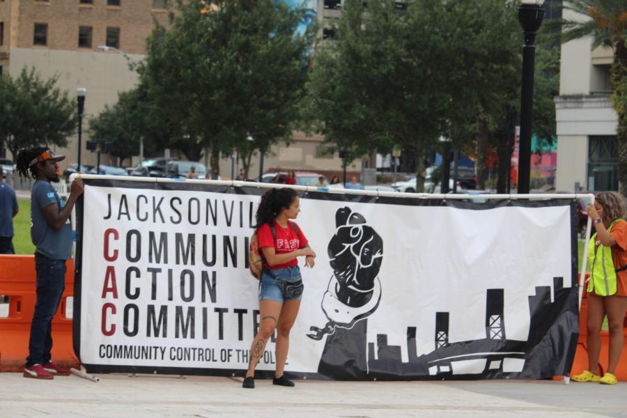 Protesters hold up a large horizontal banner that reads "Jacksonville Community Action Committee, Community Control of the Police" next to a graphic of a black raised fist