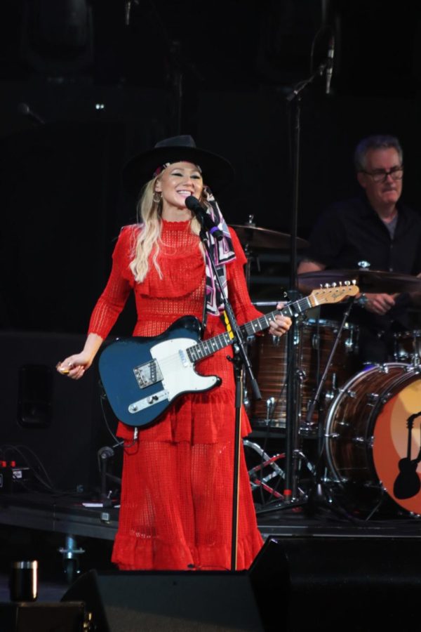 Jewel smiles on stage in a red dress and black hat while holding a blue and white guitar