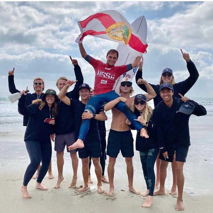 The UNF Surf Team stands on the beach waving the Florida flag