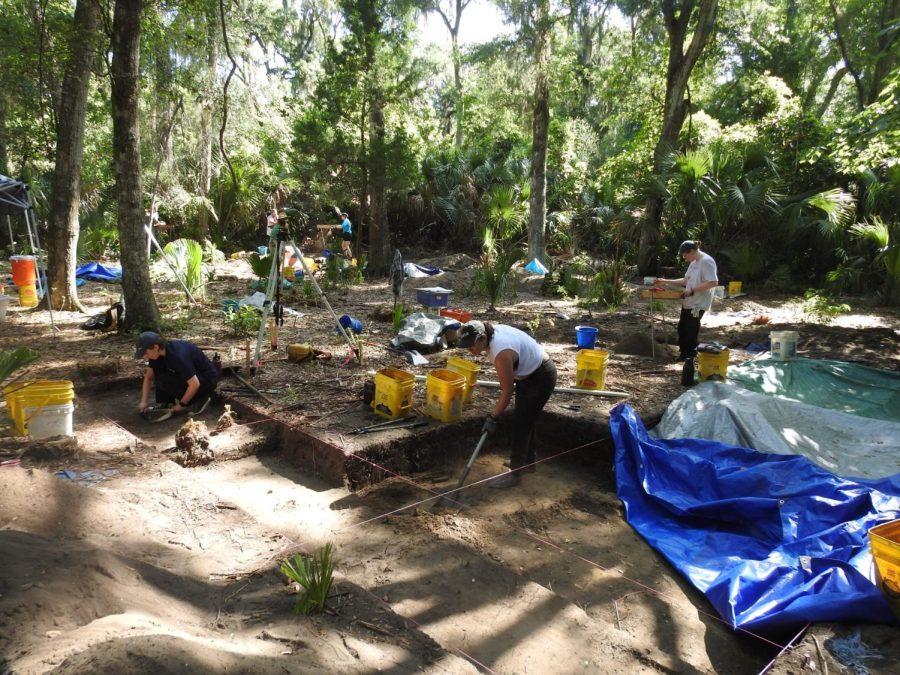 Students in masks and gloves work to uncover the trench more with yellow buckets and other tools