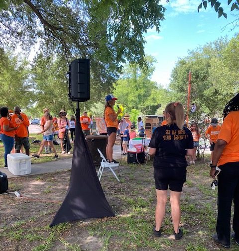 A volunteer stands on a chair with a microphone talking to others at a Wear Orange clean-up event