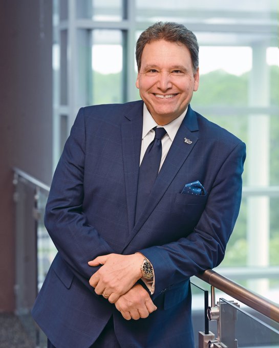 Moez Limayem leans against a railing, wearing a blue suit and tie, smiling for a photo