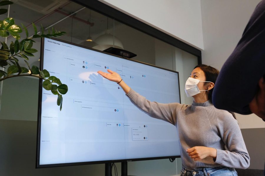 A woman wearing a white mask gestures at a TV while giving a powerpoint presentation