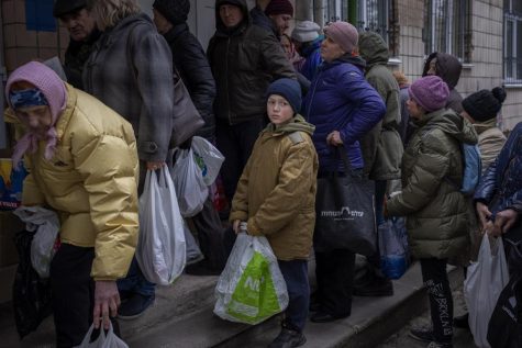 Men, women and children stand in line with plastic bags, waiting in line for food