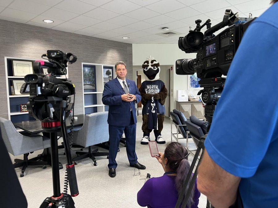 Limayem stands facing two cameras on the left and right. He is wearing a blue suit and Ozzie the Osprey stands in the background