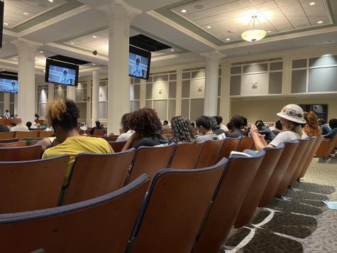 TVs hang from the ceiling with white pillars around the room. People sit in brown, cushioned chairs while the city council sits at the front of the room