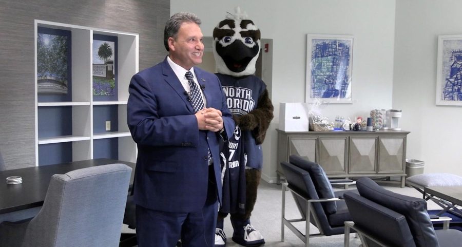 Liameyem stands. facing the camera smiling with his hands clasped. He is wearing a blue suit. Ozzie the Osprey stands behind Limayem