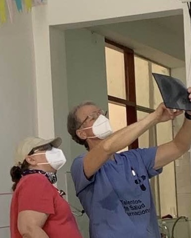 Teresa Steele, wearing a red shirt and white mask, stands next to her husband, wearing a blue shirt and glasses and a white mask, as they look at a black X-ray