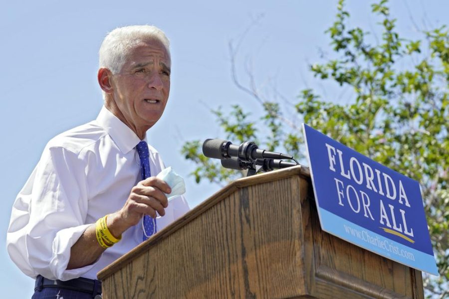 Charlie Crist talks behind a podium that has a blue sign reading "Florida For All" Crist wears a white suit, blue pants and a blue tie