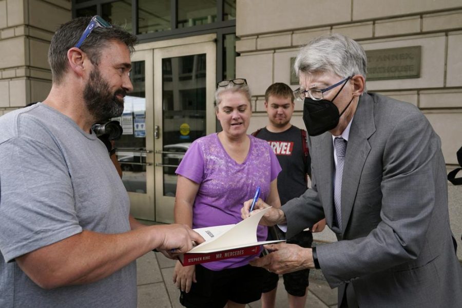 Wearing an all grey suit and black mask, Stephen King leans down, pen in hand, to autograph an open copy of a fan's book