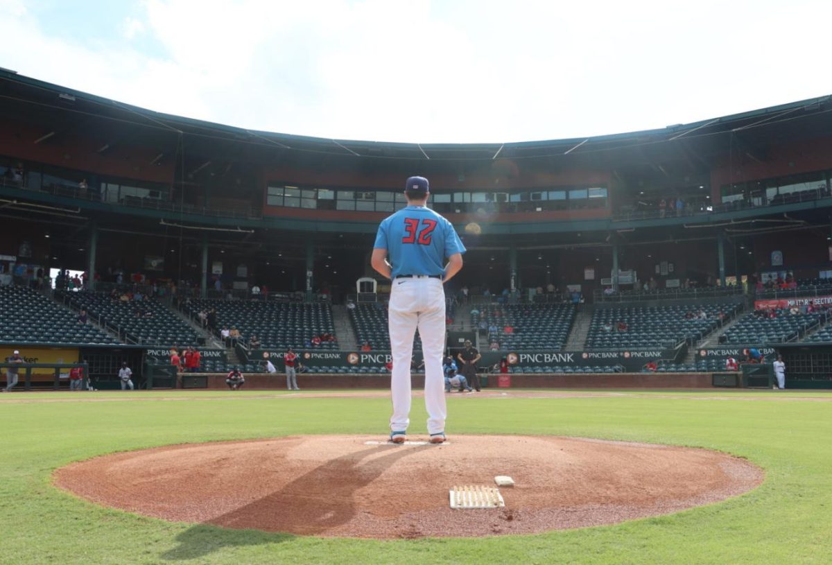 A baseball pitcher stands on the pitcher's mound, wearing white pants and a blue shirt with the number 32 on the back, facing away from the camera toward home plate.