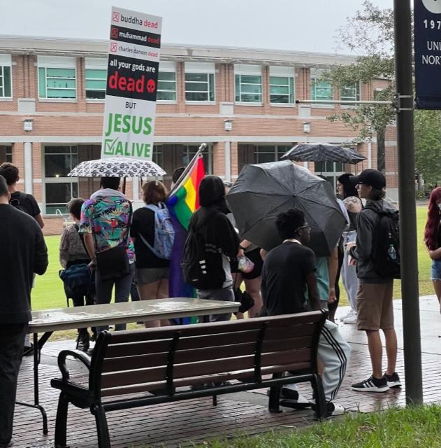 Students with umbrellas, rain coats and a pride flag gather around a preacher on the Green