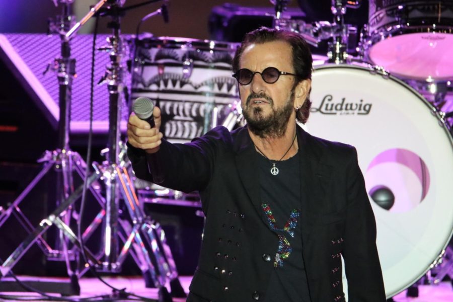 Ringo Starr holds out a microphone while wearing round black sunglasses and an all black outfit