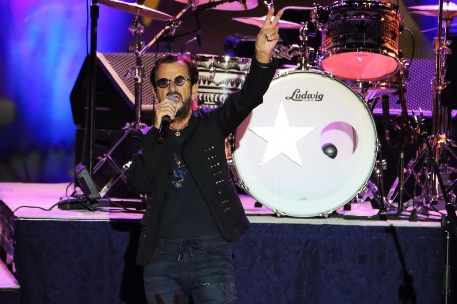 Ringo Starr holds a microphone while wearing round black sunglasses and an all black outfit
