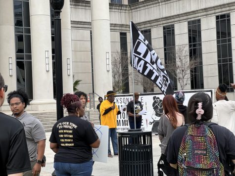 Ben Frazier stands in the center of the photo wearing a yellow shirt and black hat and holding a microphone. He is surrounded by protesters wearing various types of black and gray shirts. A "Black Lives Matter" flag waves overhead