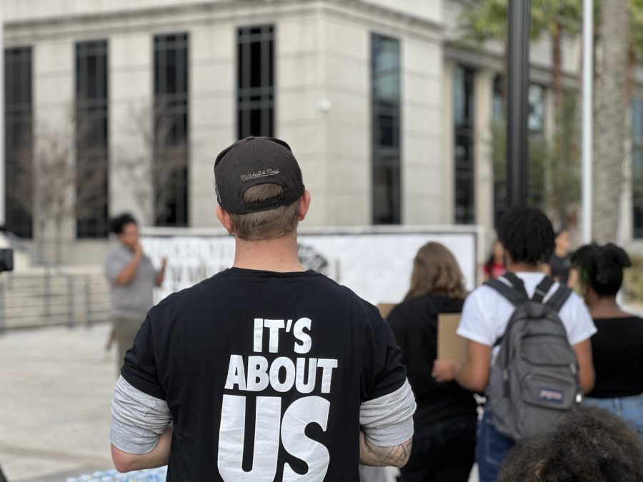 A protester faces a speaker while wearing a black hate and a black shirt with the words "It's About Us" on the back