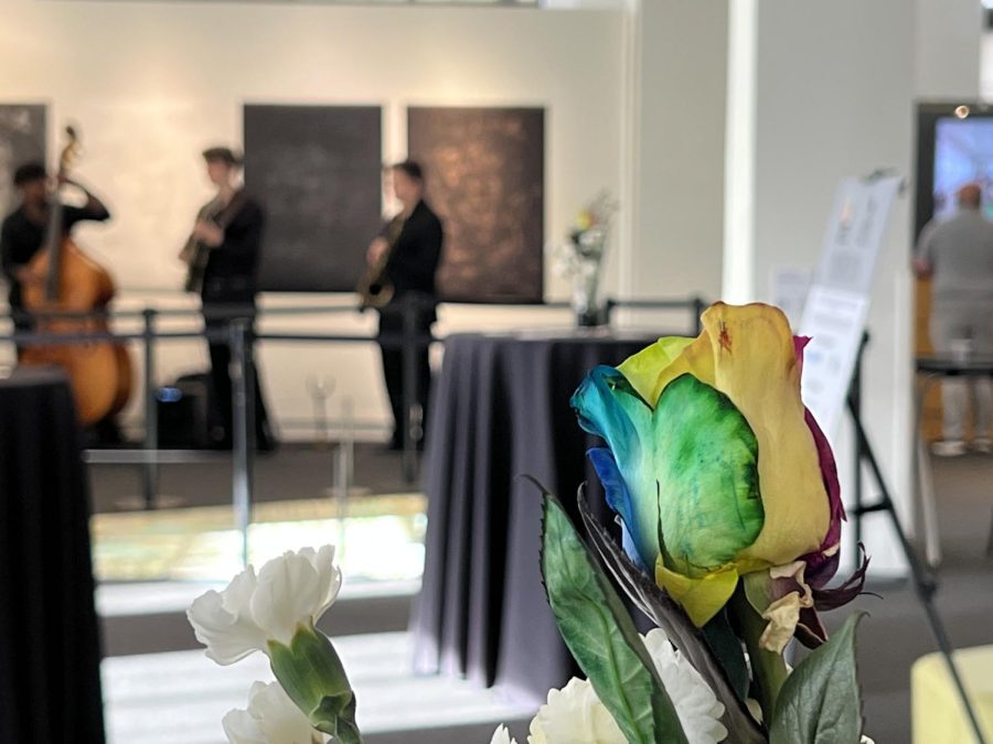 A rainbow-dyed rose in focus on the right side of the image. Three members of the small band are blurred in the back