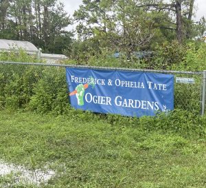 A blue sign in front of a fenced-in garden that says "Frederick & Ophelia Tate Ogier Gardens"