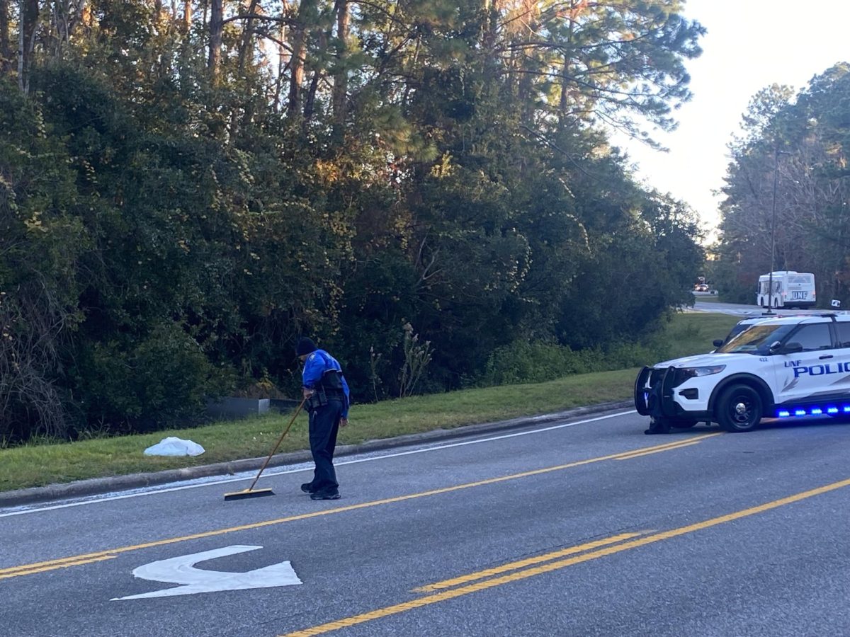 A UPD officer sweeps broken glass off the road on UNF Drive outside Tom and Betty Petway Hall on Thursday afternoon. Two police cars with lights on blocked the right lane and a police truck (not pictured) was parked on the other end of the blocked section. 