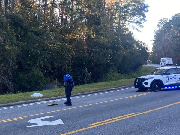 A UPD officer sweeps broken glass off the road on UNF Drive outside Tom and Betty Petway Hall on Thursday afternoon. Two police cars with lights on blocked the right lane and a police truck (not pictured) was parked on the other end of the blocked section. 