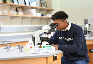 A student looks through a microscope in a science lab. He has brown hair and is wearing a blue sweatshirt.