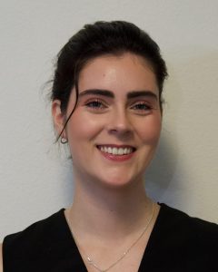 Rachel has dark brown hair and is wearing it in a bun, smiling for a photo in a black shirt.