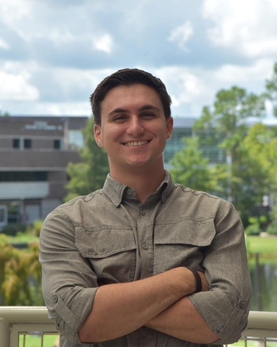 David is smiling, wearing a grey button-down shirt with his arms crossed