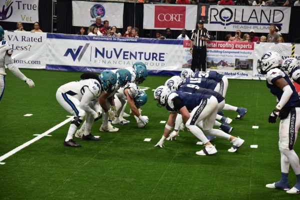 Two lines of football teams get ready for their next play on the field, one team wears UNF Osprey helmets and dark blue jerseys, the other wears white jerseys and teal helmets.