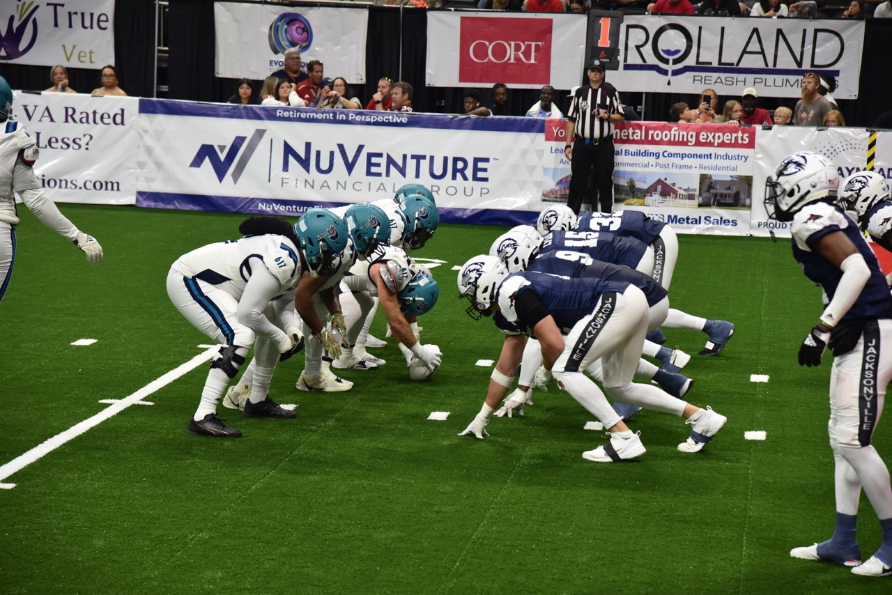 Two lines of football teams get ready for their next play on the field, one team wears UNF Osprey helmets and dark blue jerseys, the other wears white jerseys and teal helmets.