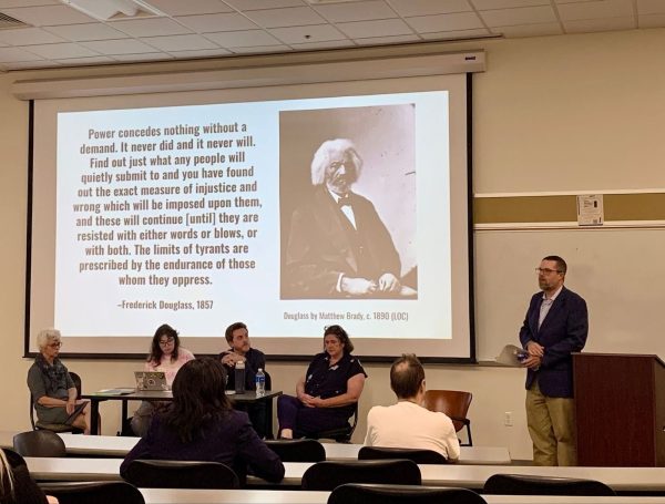 Panel discussion in a classroom with five seated speakers and one standing presenter; a large screen behind them displays a quote by Frederick Douglass and a historical photo of him.