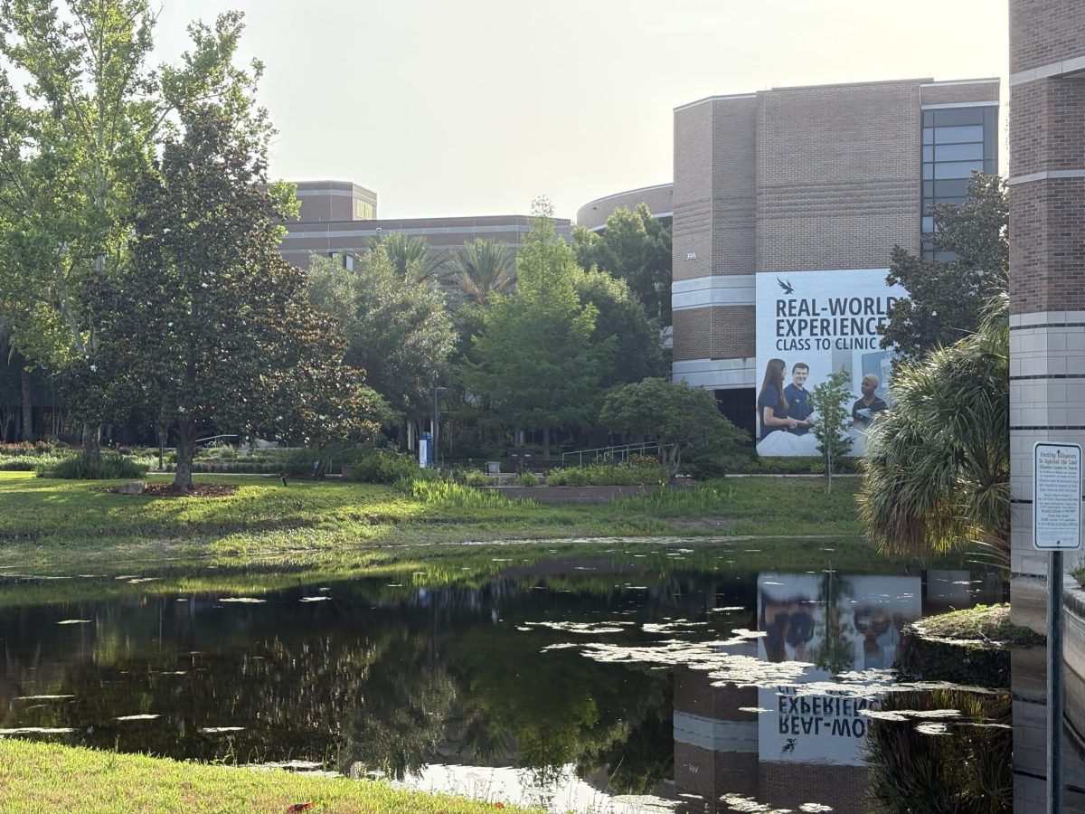A lake in front of a UNF building surrounded by green grass and trees.