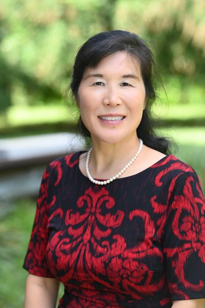 Dr. Mei Zhao is wearing a black and red blouse, a white pearl necklace and is smiling.
