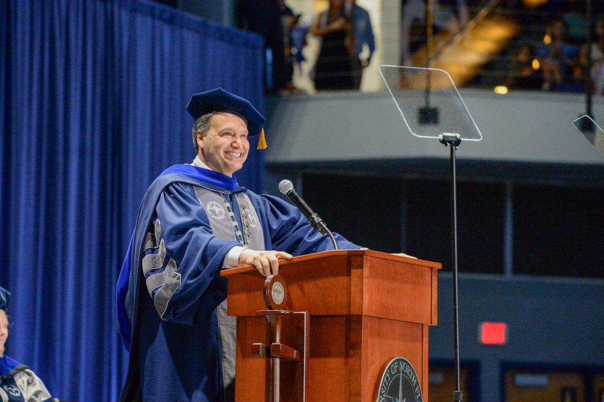 UNF President Moez Limayem speaks at a brown podium at Spring 2025 commencement. He is wearing a dark blue robe and black graduate's cap and smiling.