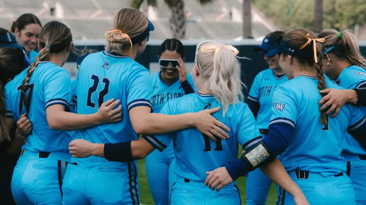 The UNF Softball team in a huddle, wearing bright blue uniforms and smiling
