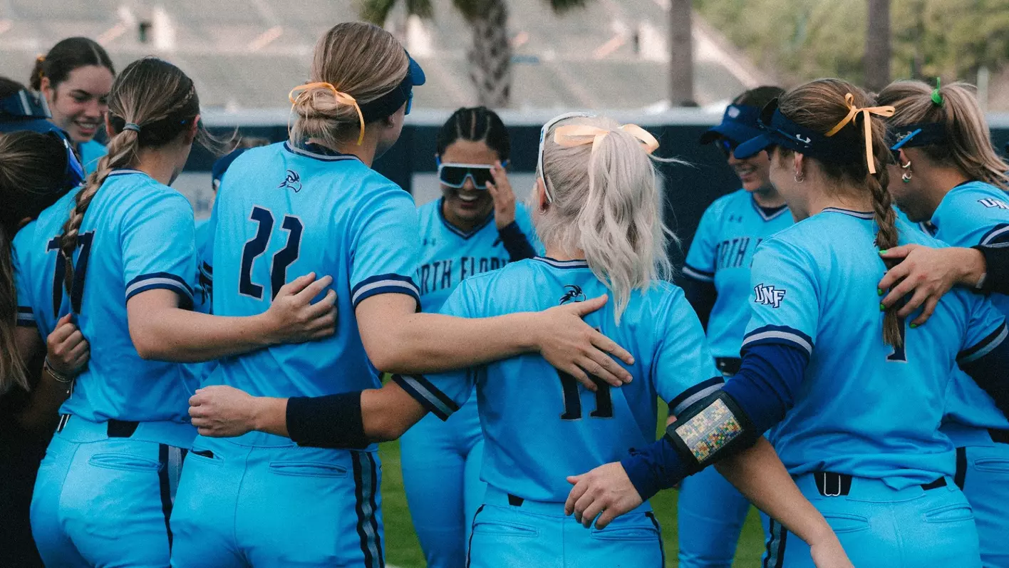 The UNF Softball team in a huddle, wearing bright blue uniforms and smiling