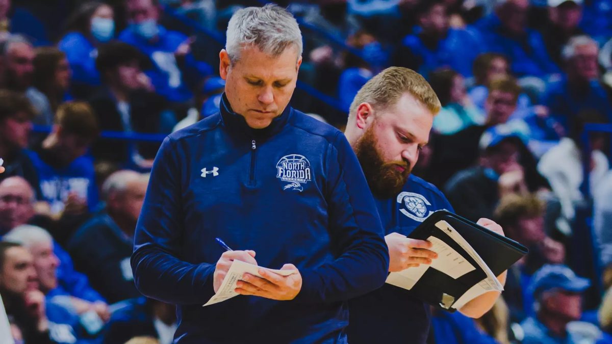 New Men's Basketball Head Coach Bobby Kennen takes notes at a basketball game in a blue jacket