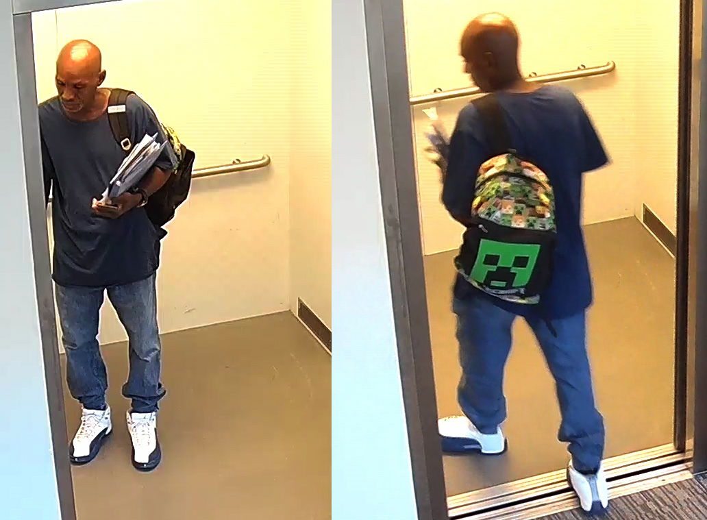 A man walks into an elevator, wearing a black shirt, blue jeans, and white shoes. He is carrying papers and has a Minecraft backpack on.