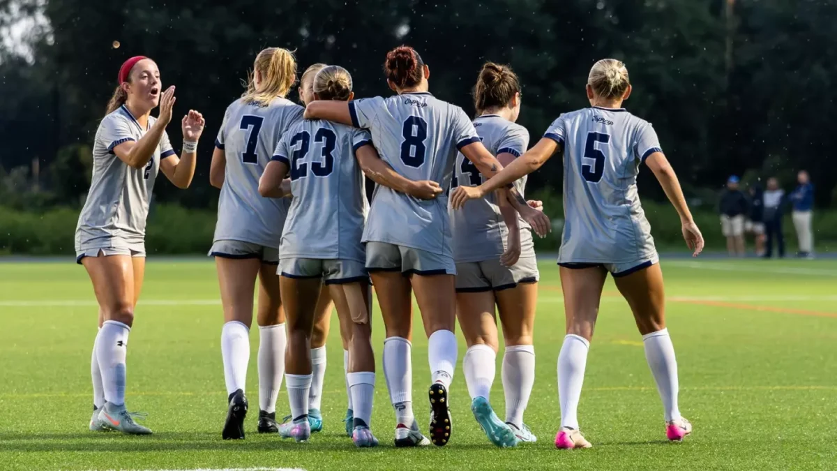 Six female soccer players embrace side-by-side, with their backs turned away from the camera. They wear all grey jerseys.