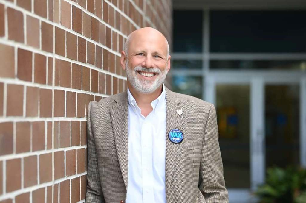 John white is smiling, wearing a tan suit jacket and white shirt, standing next to a red brick wall.