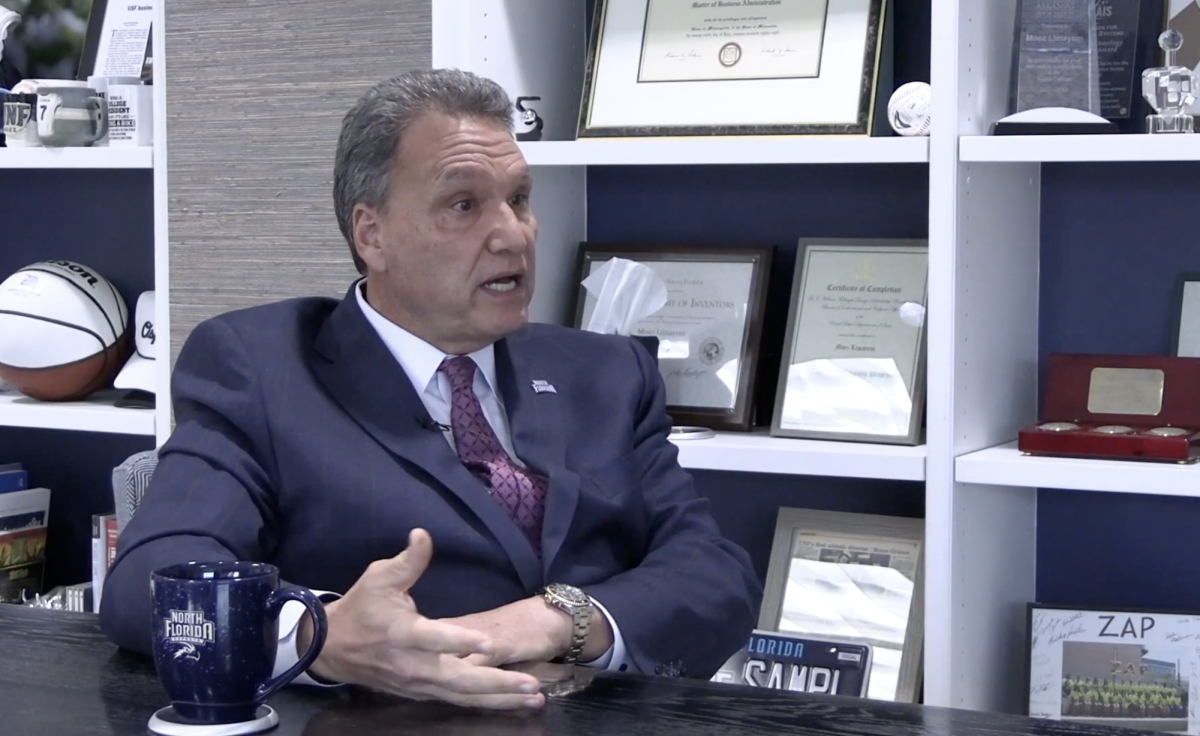 UNF President Limayem speaks, he's sitting in his office in front of a white book shelf wearing a dark blue suit.