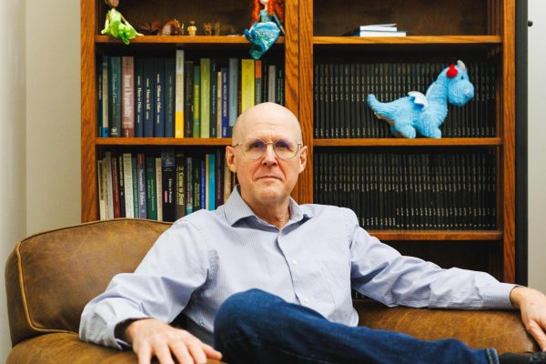 George Rainbolt sits in a brown chair in front of a bookshelf. He's smiling and wearing a blue shirt and jeans