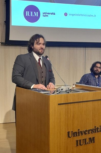 McElroy speaks at an academic conference, wearing a suit and smiling behind a wooden podium