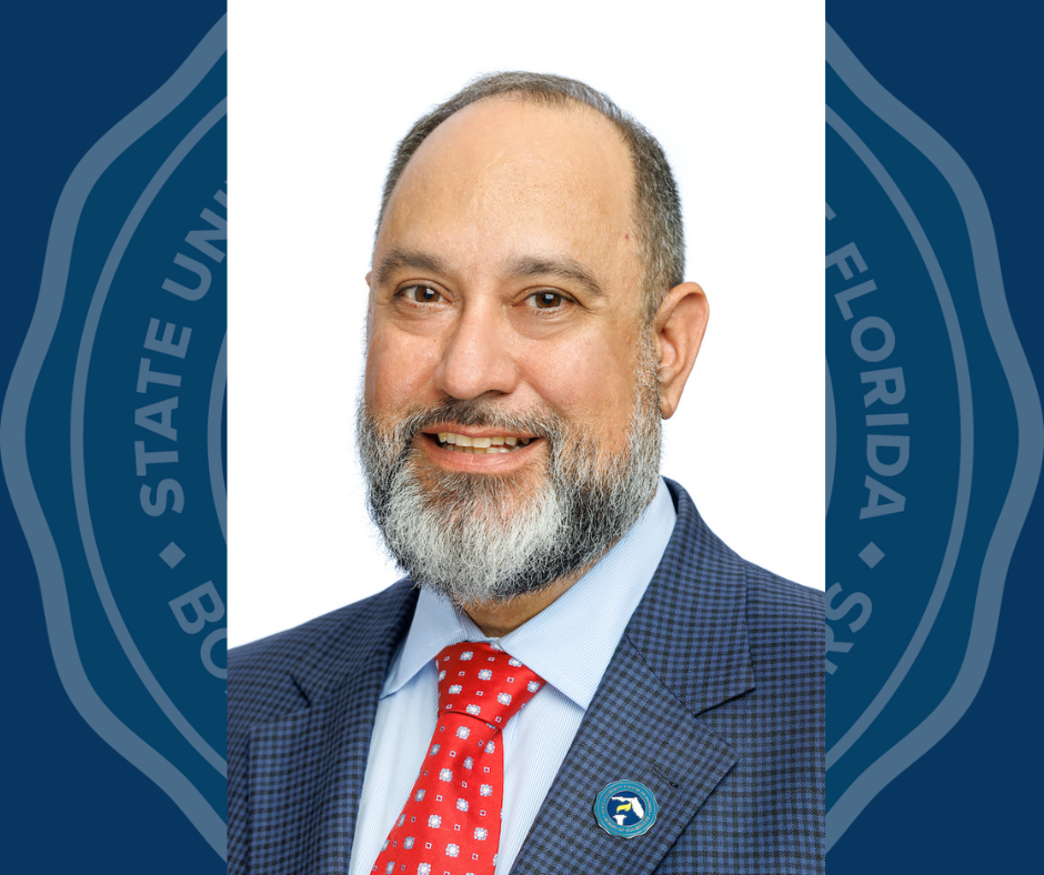 Florida SUS Chancellor Ray Rodrigues smiles. wearing a blue suit with a red tie, he has a beard and is balding