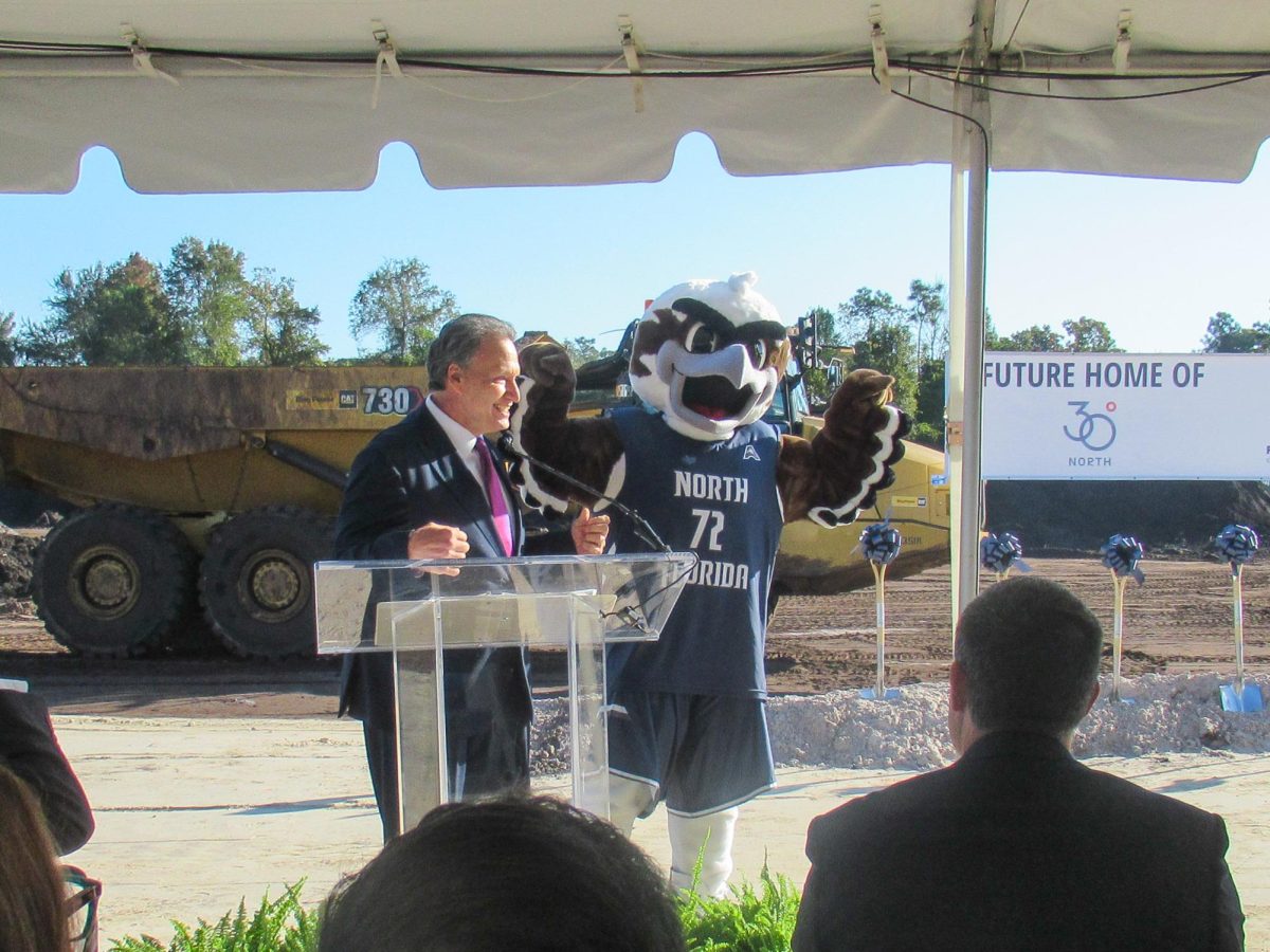 A man in a blue suit stands at the front of a glass podium, next to a man wearing a bird mascot costume.