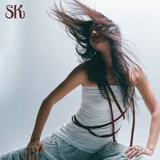 A brunette girl wearing a white shirt flips her hair. She's sitting in front of a white background and has a red ribbon draped on her with the letters "SK" in red written in the top left corner.
