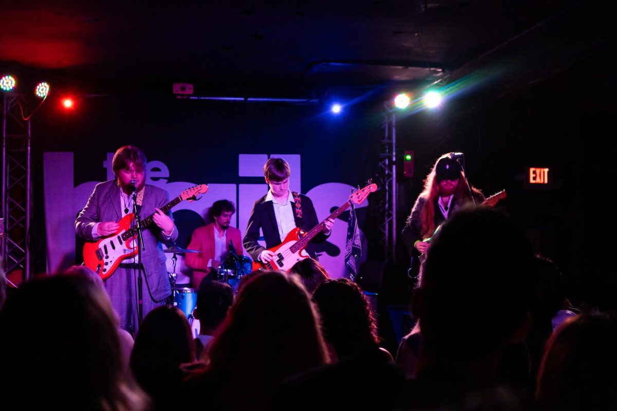 Three guitarists and one drummer play on stage in purple lighting.